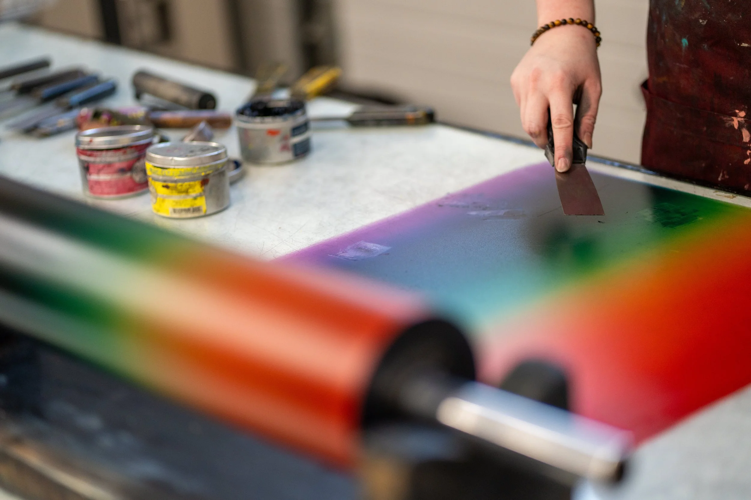 Close-up of a person wearing a bracelet, holding a palette knife, with ink cans and tools on a table.