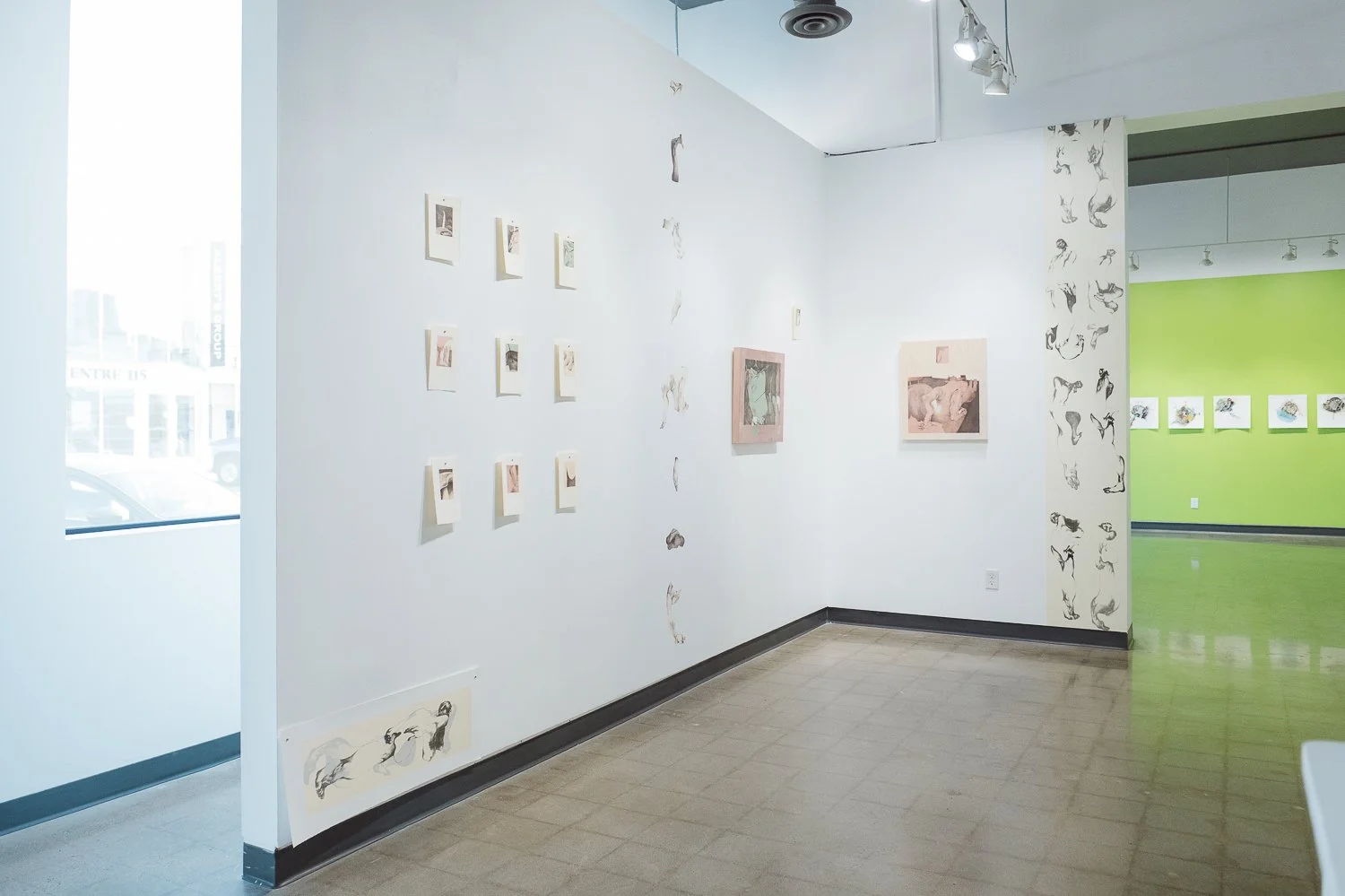 An art gallery with white walls displaying various small prints and drawings, a window letting in natural light, and a corridor with a green wall featuring additional artwork.