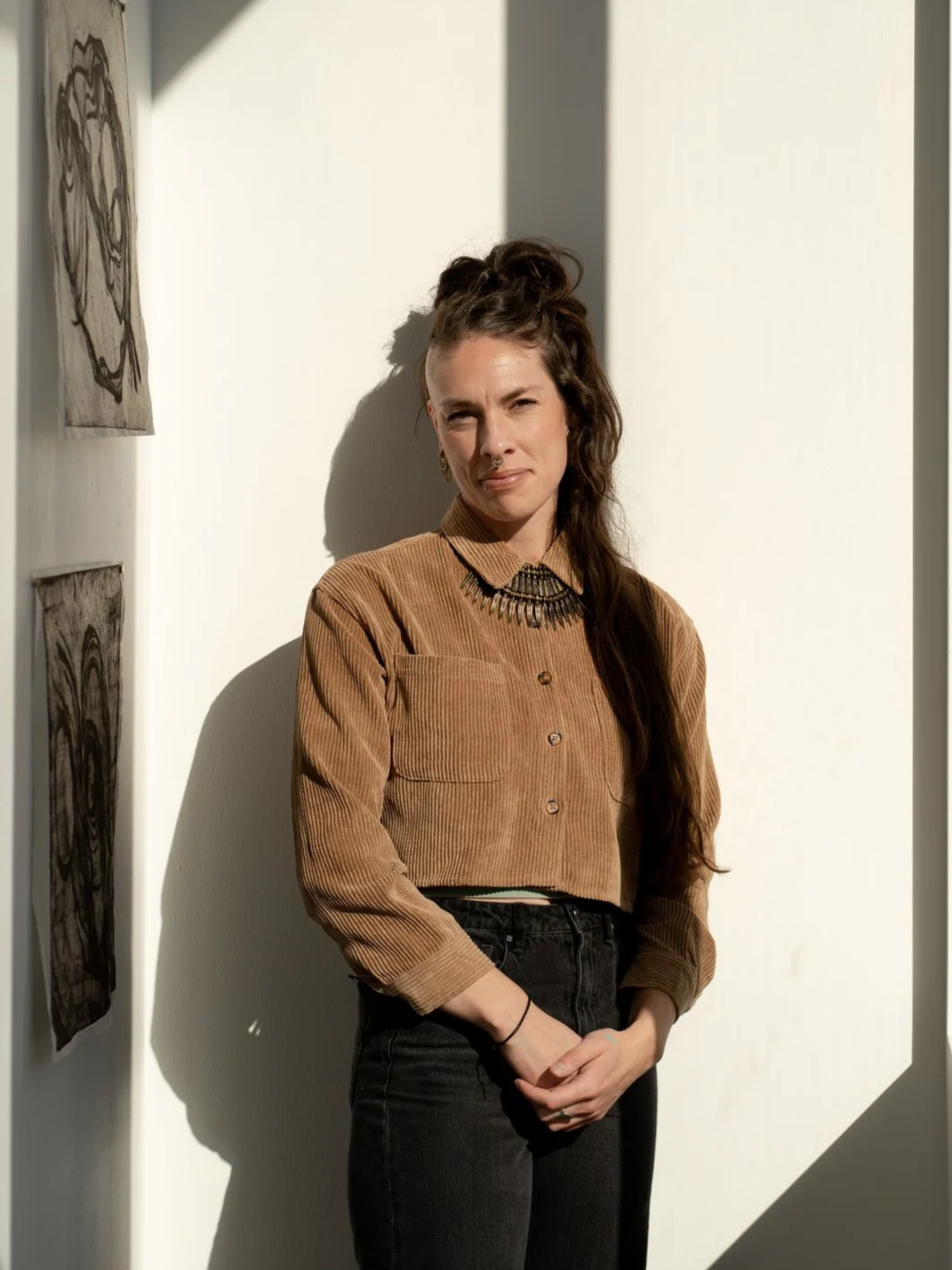 A young woman with long dark hair styled in a half-up, half-down hairstyle, standing against a white wall with sunlight casting shadows. She wears a brown corduroy button-up shirt, black high-waisted jeans, and a statement necklace. She has a neutral expression with a slight smile.