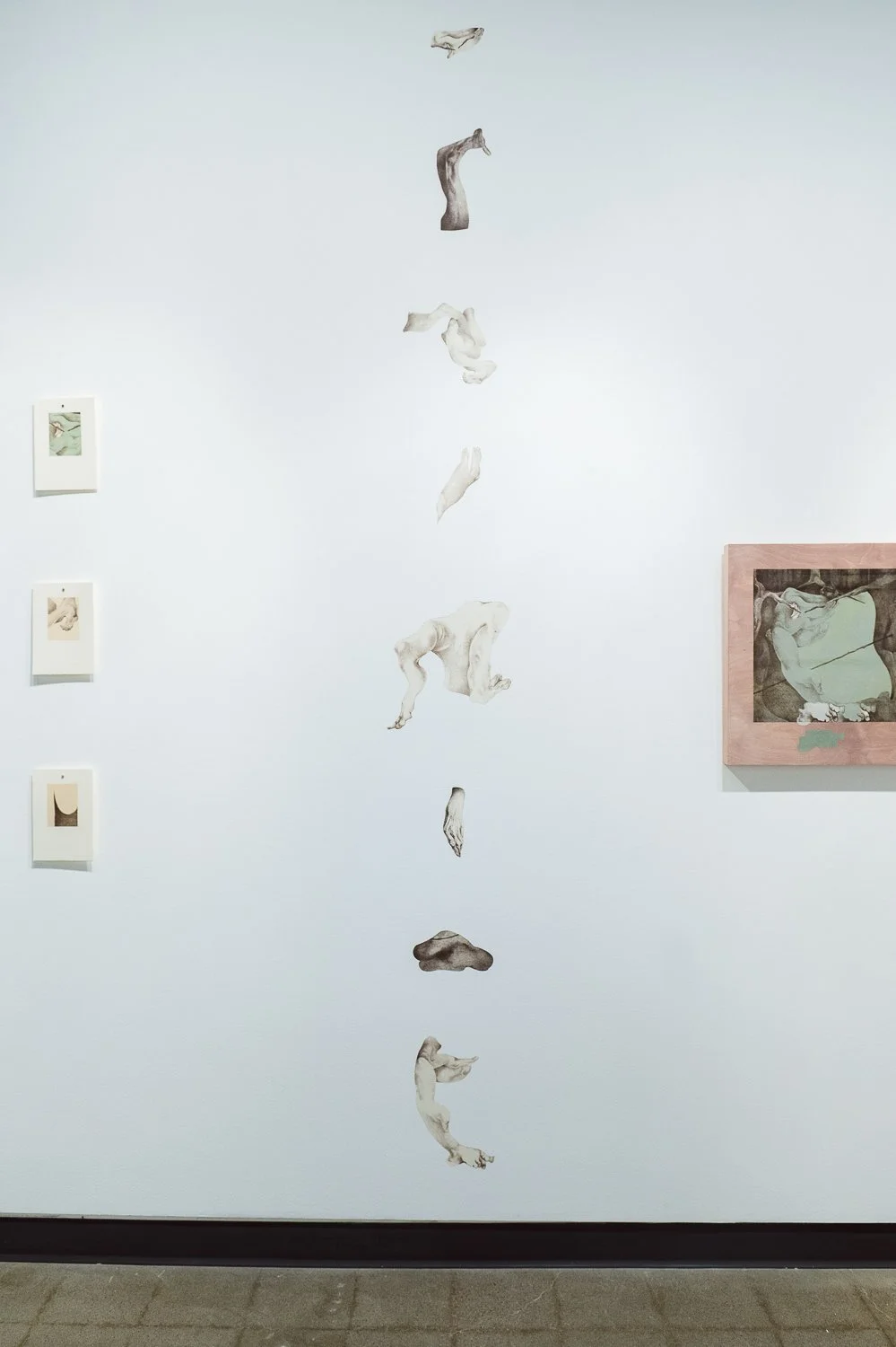 A white gallery wall with a vertical arrangement of paper cutout images of human body parts, including arms, hands, and feet, along with framed artworks on the left and right.