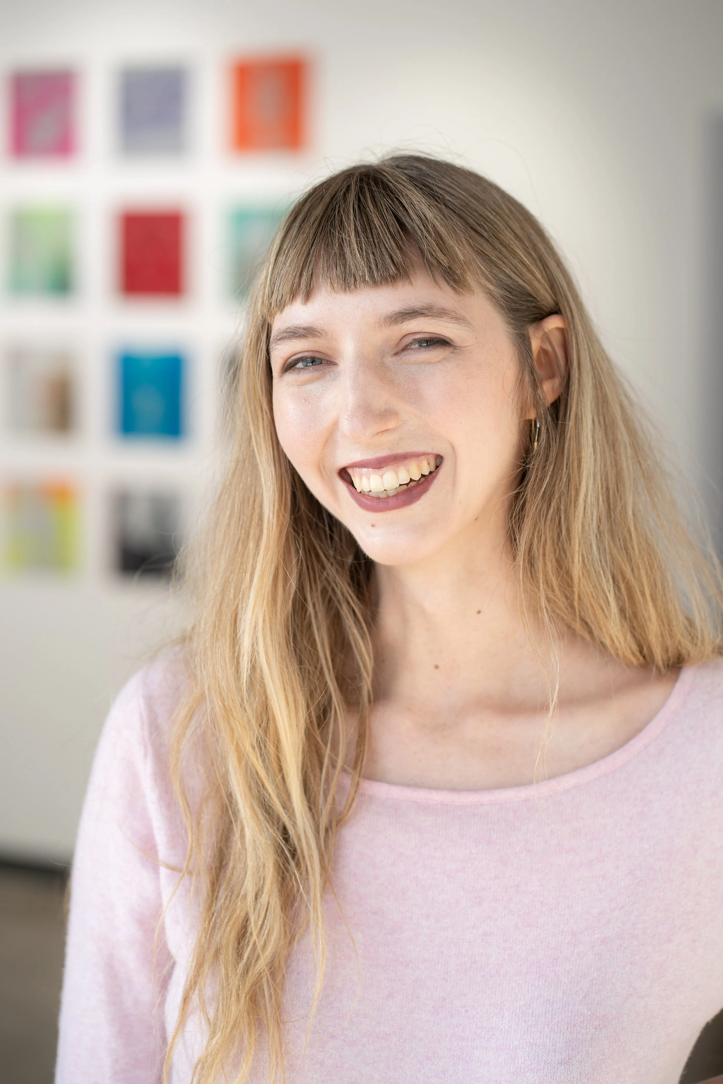 Close-up of a woman with long blonde hair, smiling and wearing a light pink top, with a blurred colorful artwork on the wall behind her.