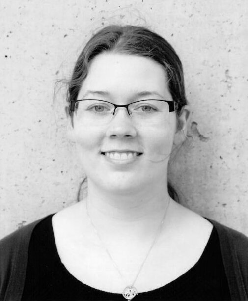 A woman with glasses and a necklace, smiling, standing in front of a concrete wall.