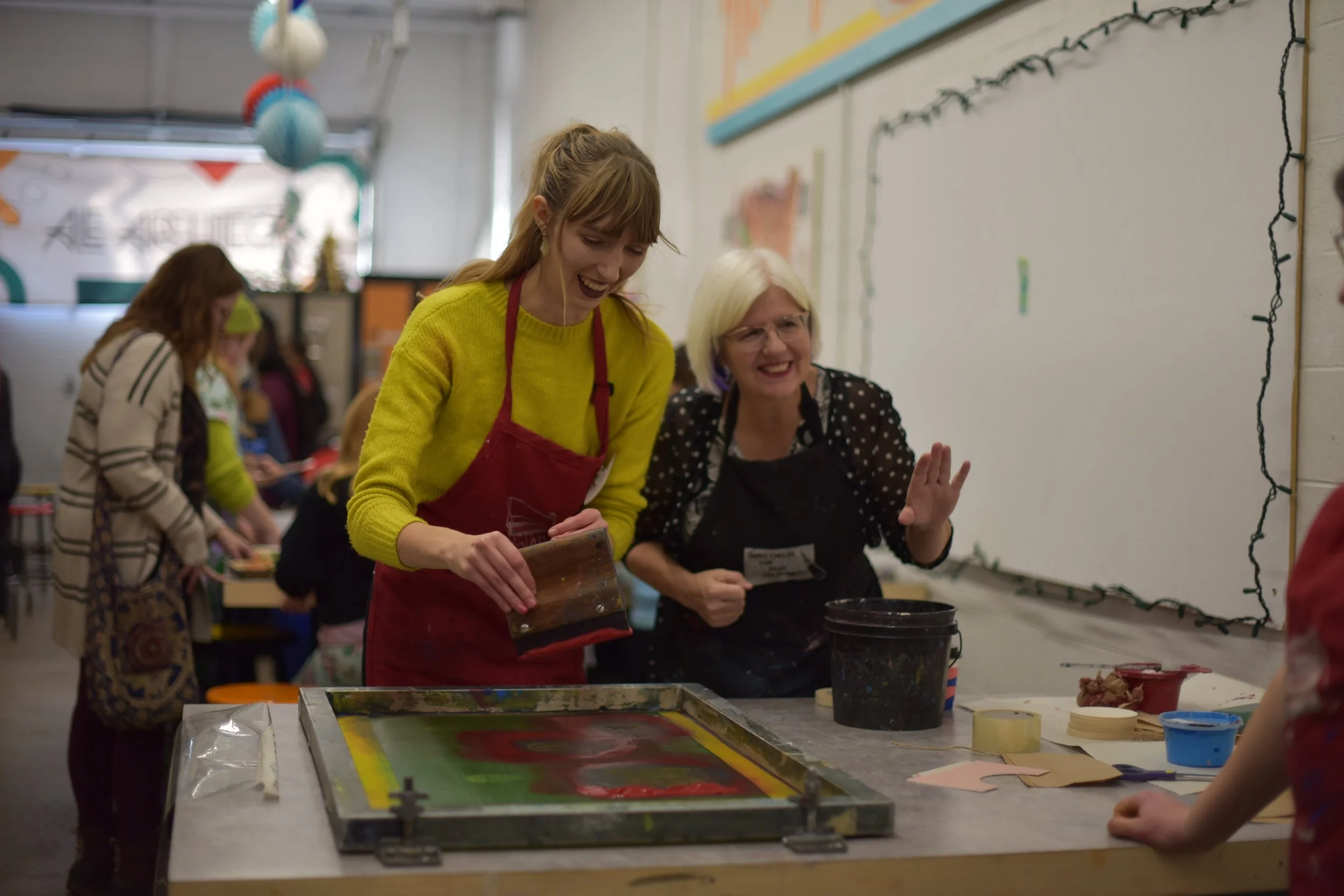 People participating in an art activity at a classroom table, with a woman in a red apron and yellow sweater smiling and looking at her phone, surrounded by others engaged in artwork.