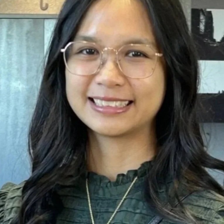 A young woman with long black hair, wearing glasses, and smiling in an indoor setting.