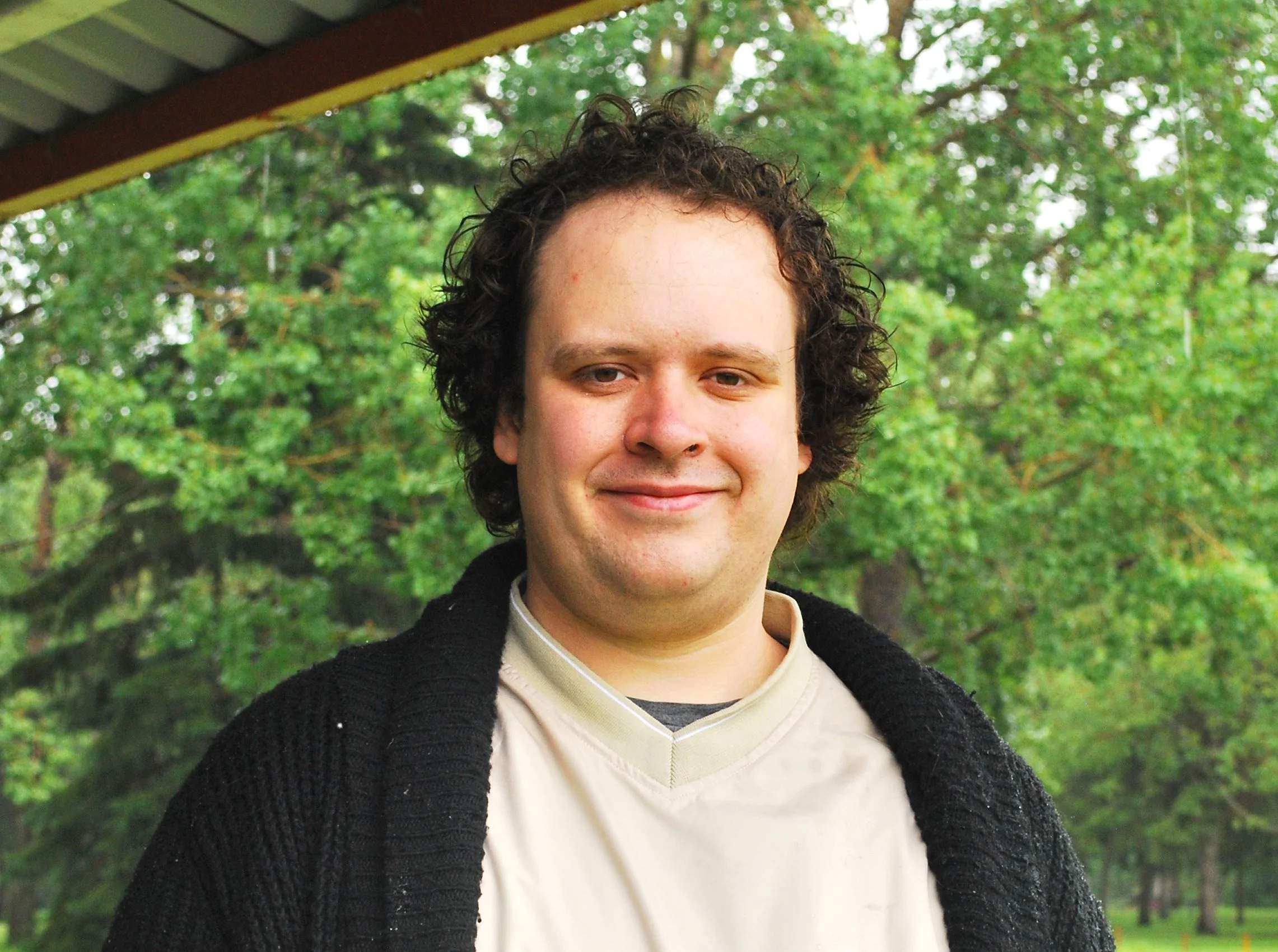 Close-up of a smiling person with curly hair wearing a cream-colored shirt and a black sweater, standing outdoors in front of green trees.