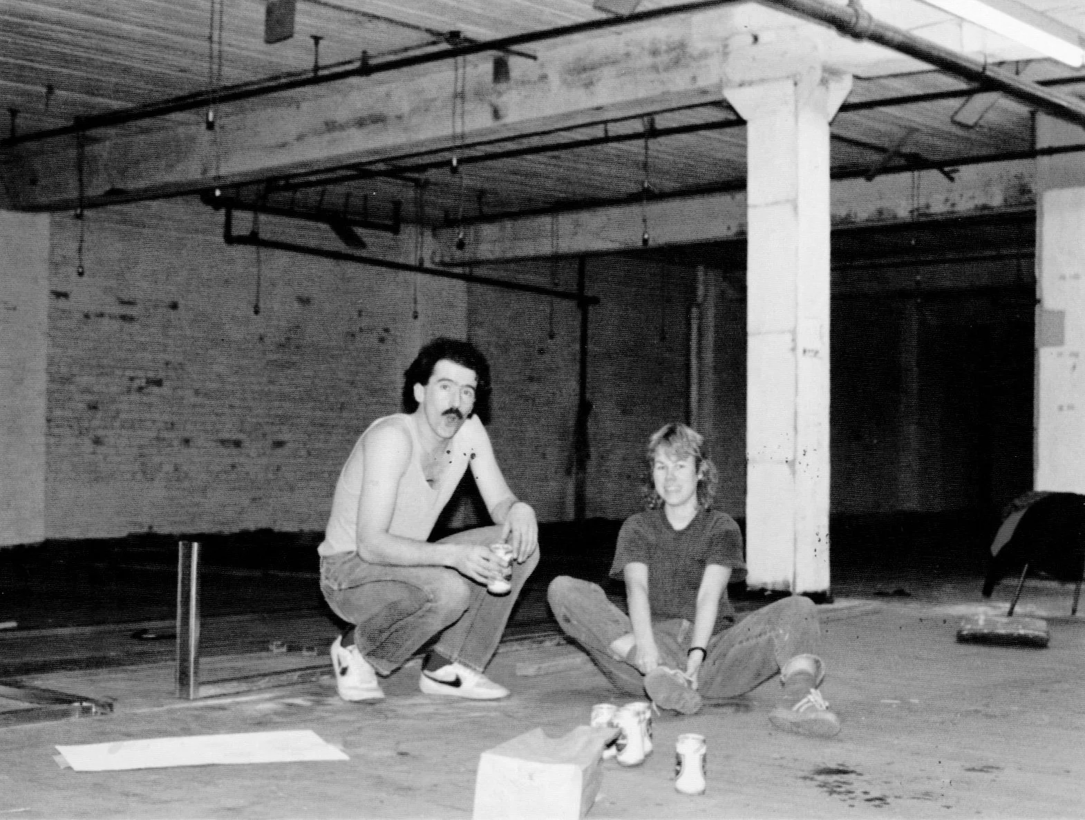 A black and white photo of two men sitting on the ground in an industrial or warehouse setting, with exposed brick walls and pipes overhead. One man has dark hair, a mustache, and is wearing a sleeveless shirt and sneakers; the other has lighter hair, glasses, and is dressed casually. They are surrounded by various objects, including cans and papers.