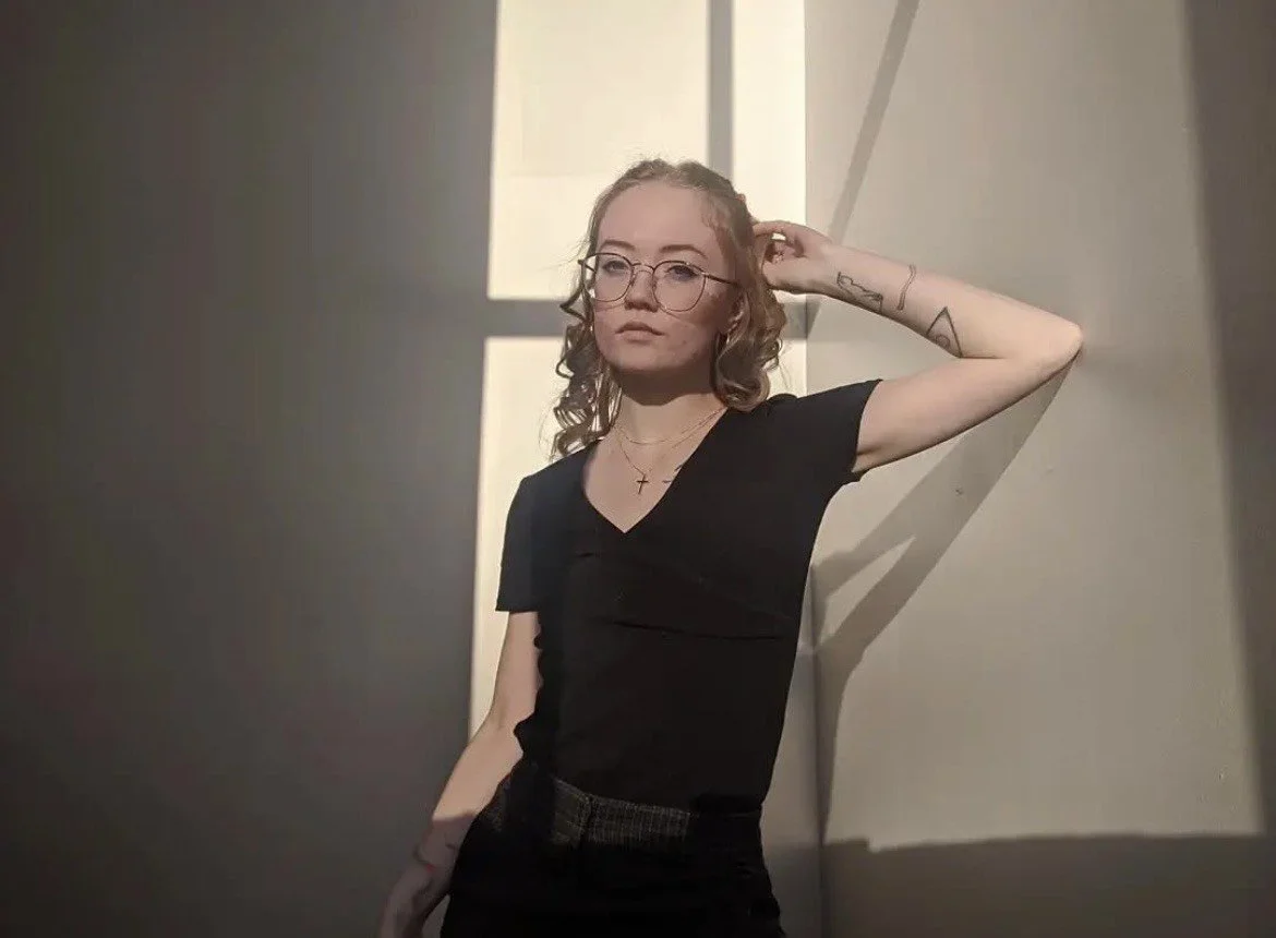 A person with curly blond hair, glasses, and tattoos on their arm, standing in a room with sunlight casting shadows on the wall.