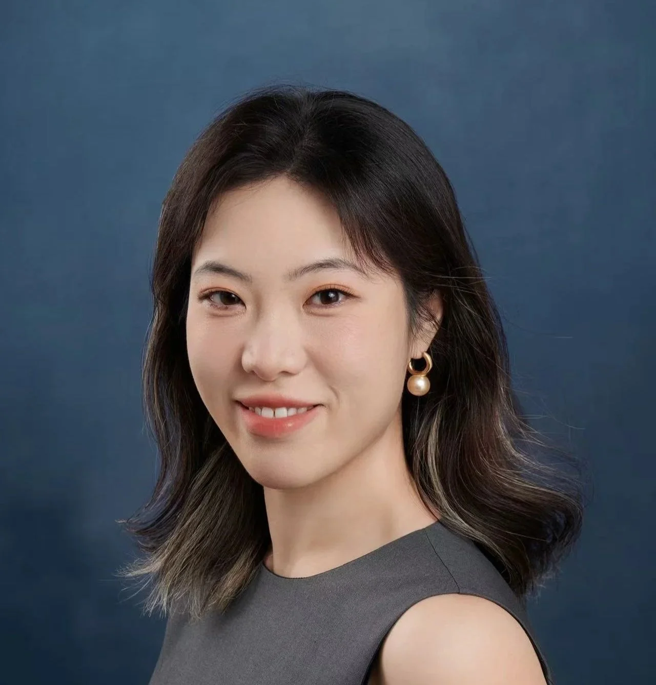 Portrait of a woman with shoulder-length dark hair, wearing pearl earrings and a sleeveless gray top, smiling at the camera against a dark blue background.