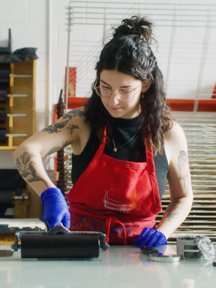 A woman with tattoos, wearing glasses, a black sleeveless top, a red apron, and blue gloves, using a brayer to print on a glass surface in an art studio.
