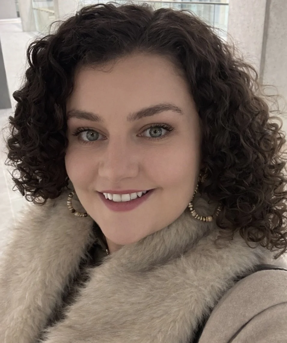 A woman with curly brown hair, wearing hoop earrings and a beige fur coat, smiling at the camera.