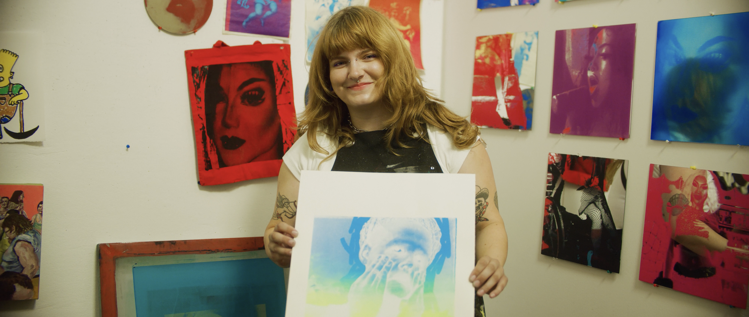 A young woman with red hair and tattoos on her arms standing in an art studio, holding a colorful print of a person in drag. The wall behind her displays various vibrant pop art prints.