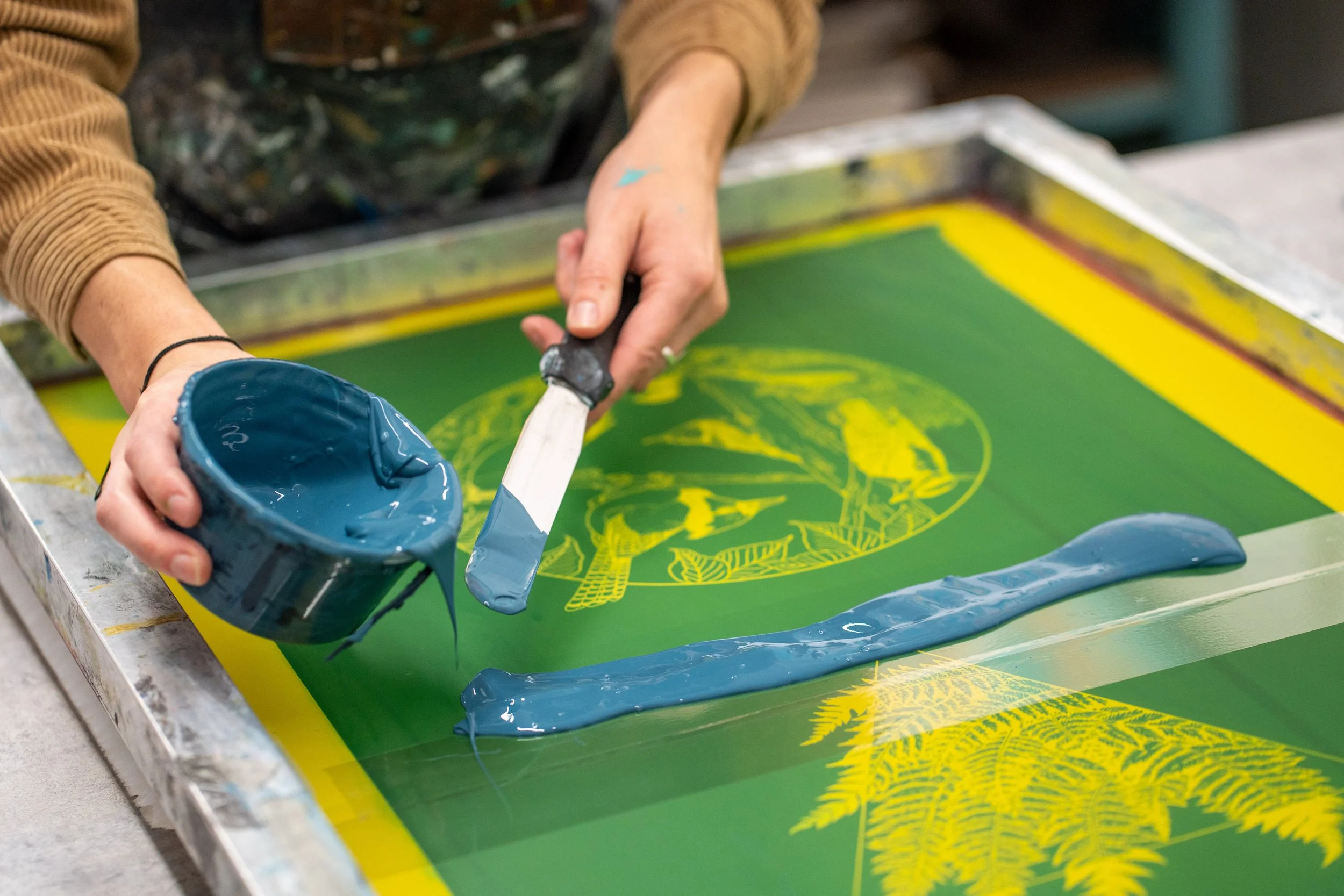 Person using a palette knife to spread blue ink onto a silkscreen printing surface.