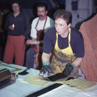 Man in an apron working with tools at a table, two men observing in the background, indoor setting.