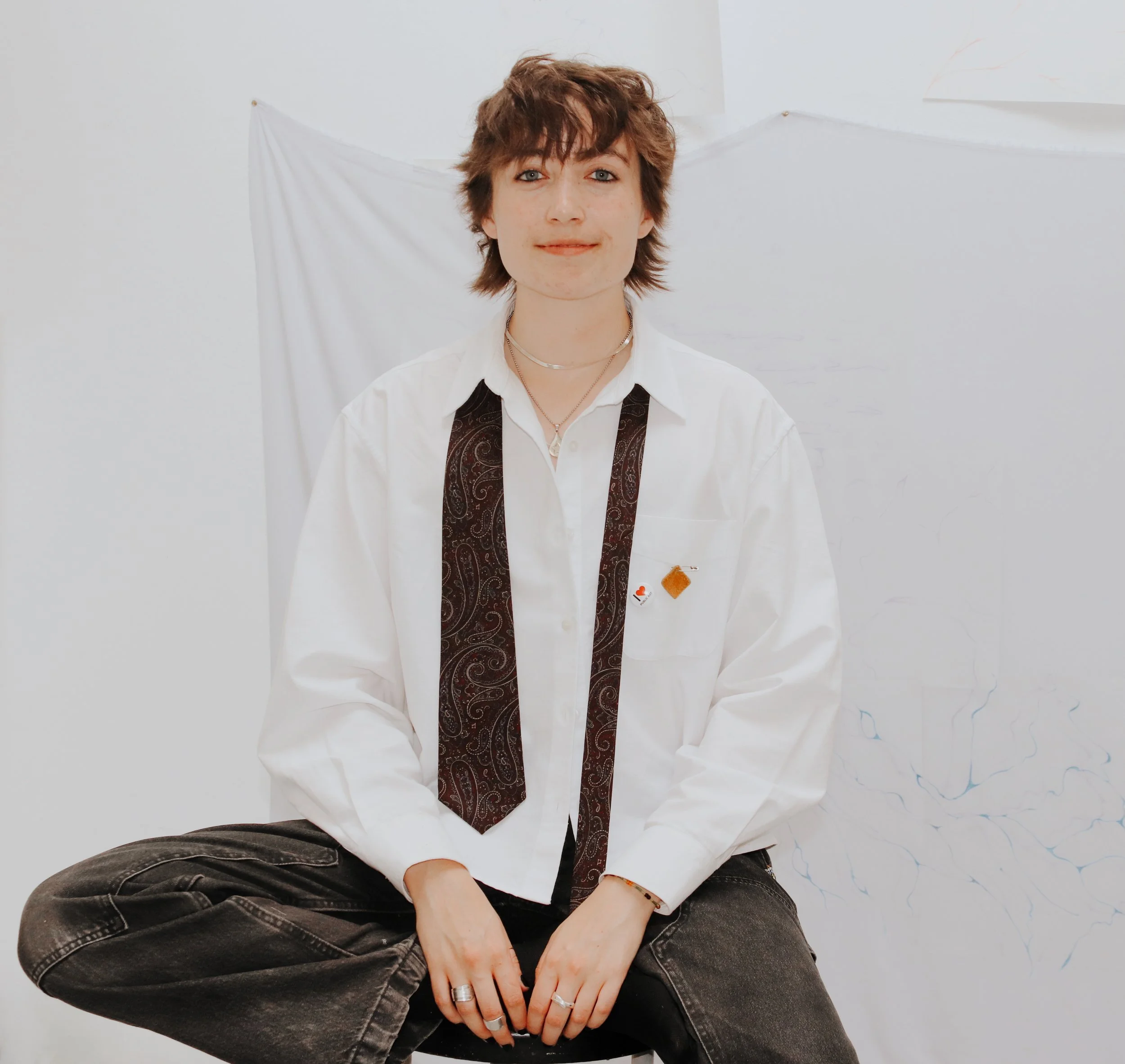 A person with wavy brown hair sitting against a white background, wearing a white shirt with a black paisley-patterned tie and black jeans, with multiple rings and a necklace visible.