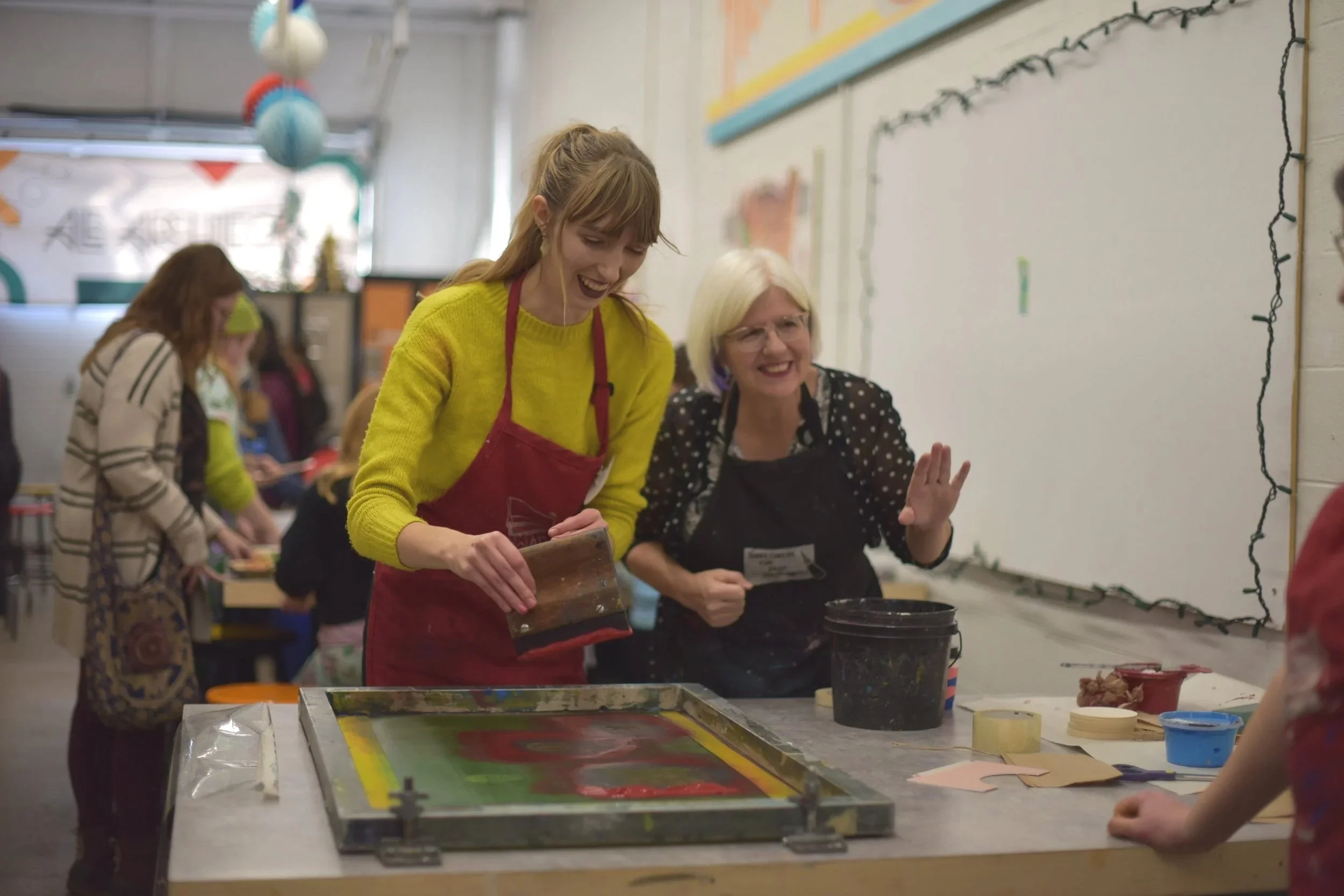 Two women are in an art studio, smiling and working on a screen printing project. They are surrounded by print supplies and other participants.