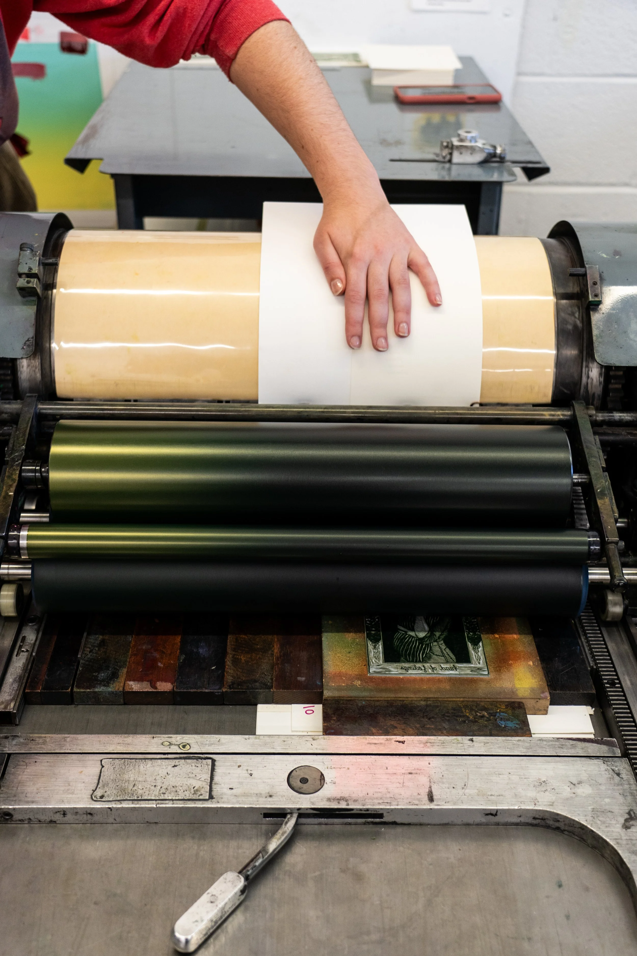 A person's hand pressing a sheet of paper against a roller in a letterpress printing machine. The machine has multiple rollers with different colours, and the background includes a work table with books and a smartphone.