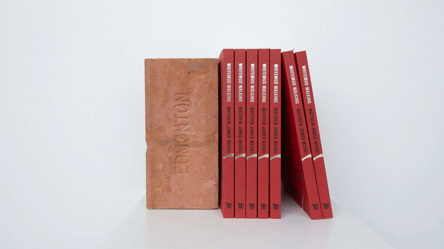 A white shelf holding a stone brick and seven red books with black and white text on the spine, titled 'Whitemud Walking' by Matthew James Weigel.