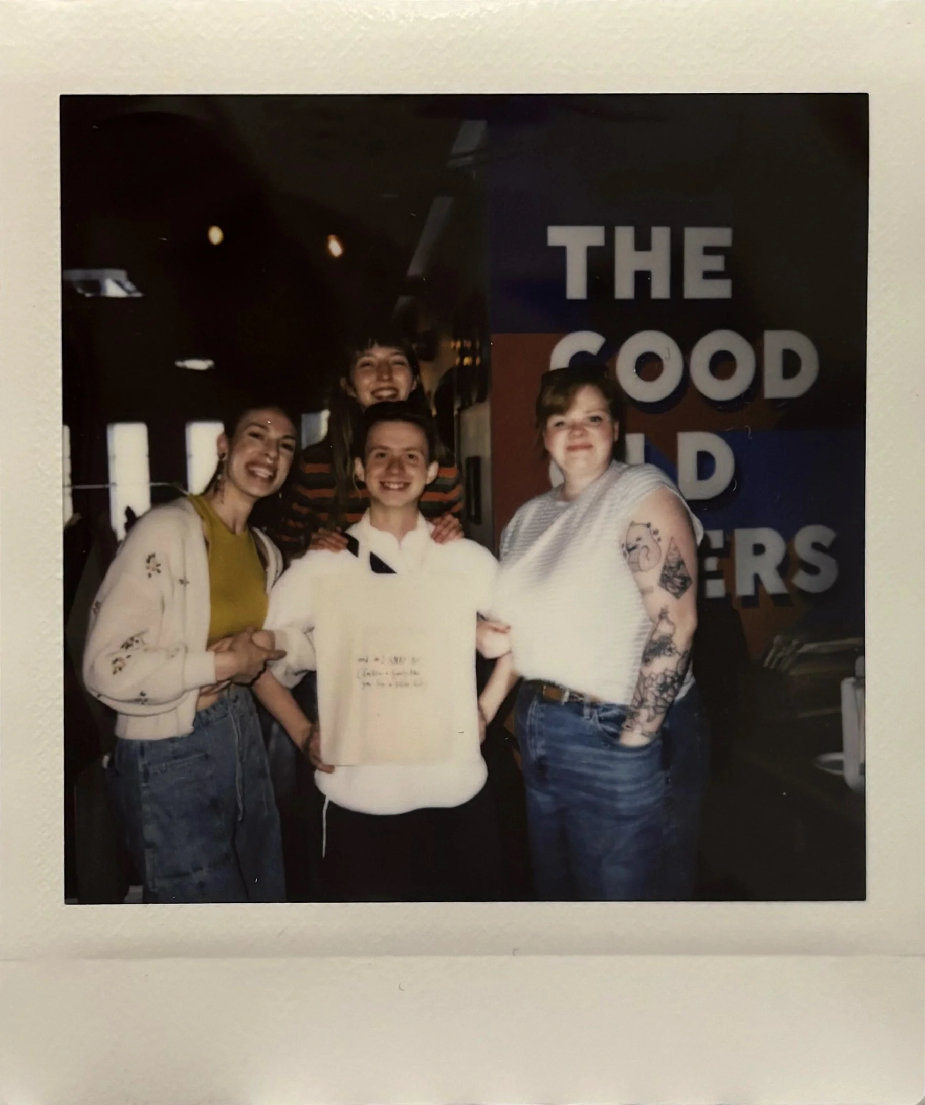 Group of four smiling people standing in front of a sign with the partially visible words 'THE GOOD' and 'R' with dark background