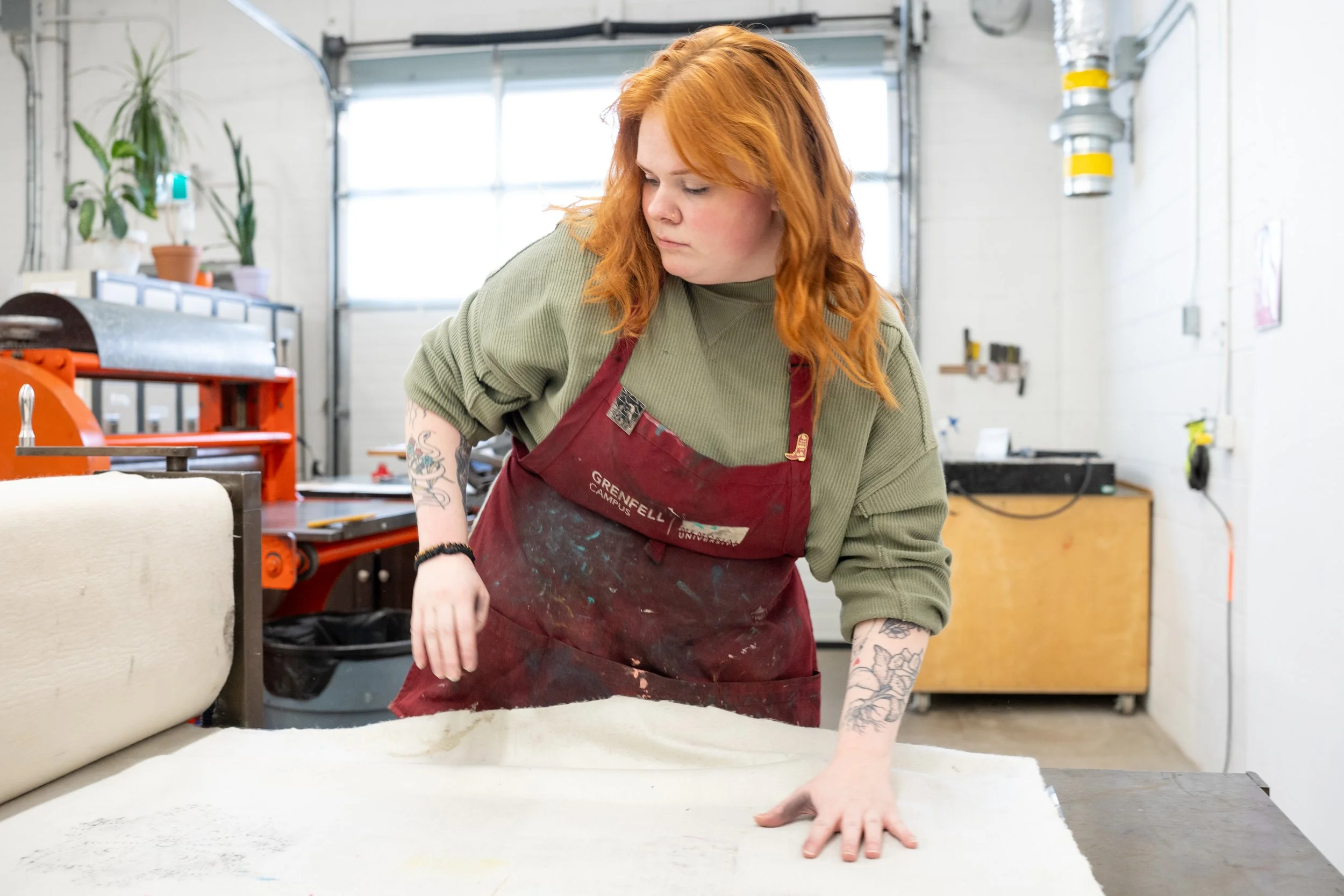 A woman with red hair and tattoos in a green shirt and red apron, working in a workshop with plants and tools, pulling a sheet of fabric or paper.