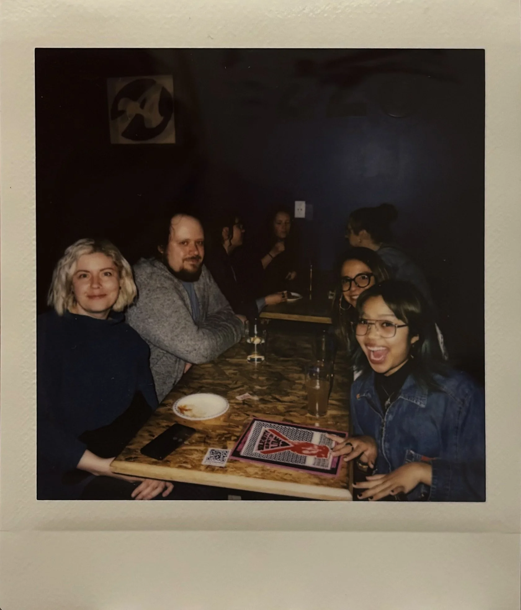 A group of six friends sitting at a wooden table in a dimly lit bar or restaurant. They are smiling and appear to be enjoying drinks and conversation. One person is holding a menu, and there are some drinks and a plate of nachos on the table.