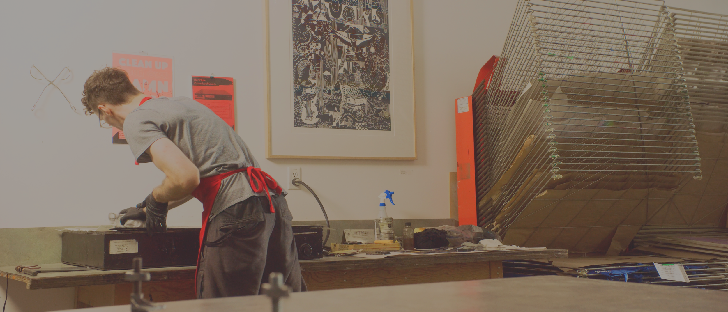 Person working in an art studio, handling a black box or container, with art posters and stacked frames on the wall and a spray bottle on the workbench.