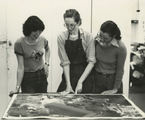 Three women looking at a large print on a table in a room with shelves and cabinets.