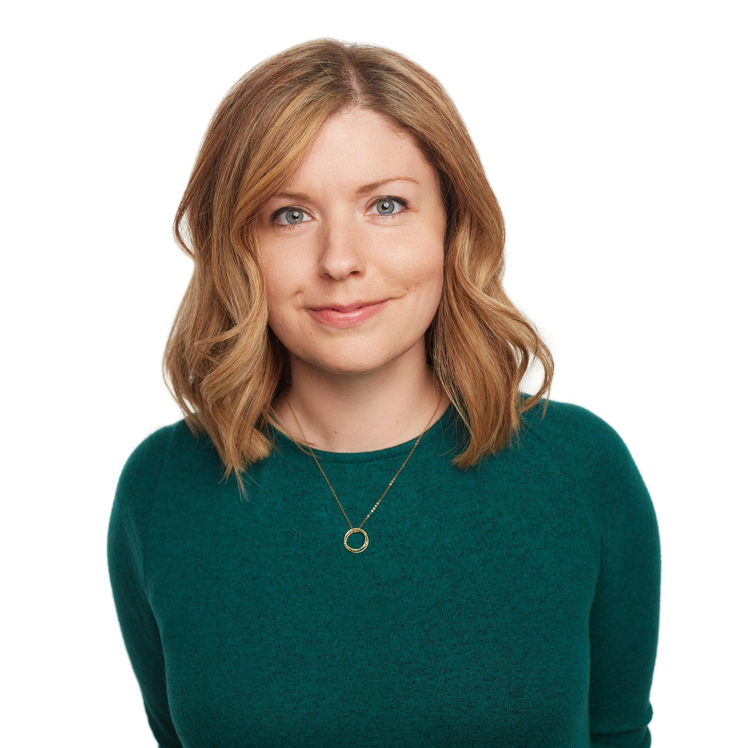 A woman with shoulder-length wavy blonde hair, wearing a dark green sweater, and a gold necklace with a circular pendant,  smiling softly against a plain white background.