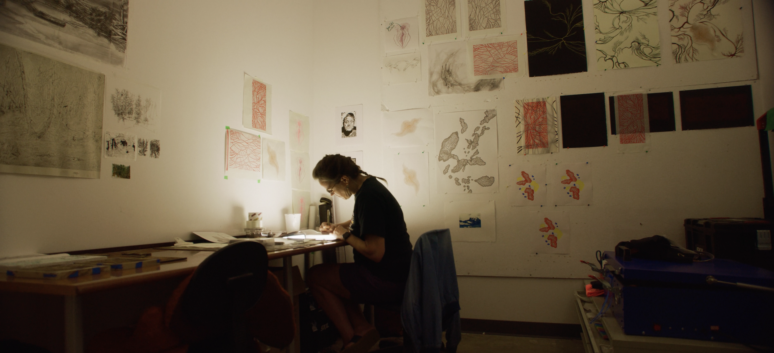 A person sitting at a cluttered desk writing in a notebook, with art and sketches hanging on the wall behind them in a dimly lit room.