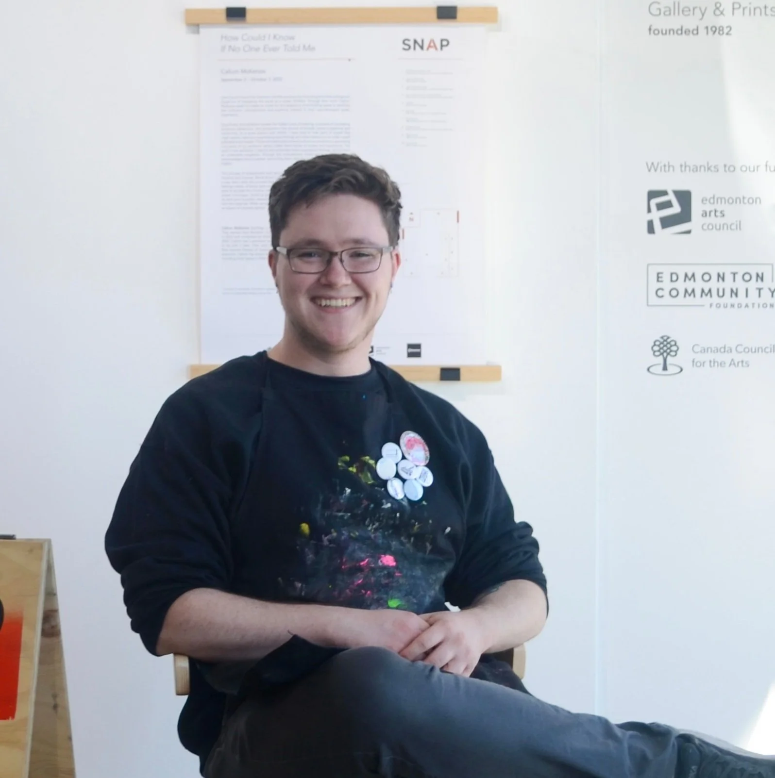 A young person with glasses, smiling, sitting on a wooden chair with his arms crossed, wearing a black sweatshirt with colorful paint stains, in front of a white wall with posters and logos.
