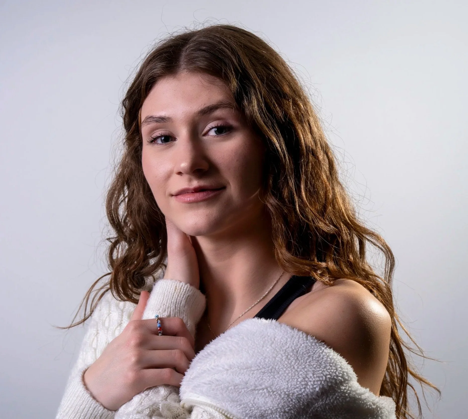 A young woman with curly brown hair, blue eyes, and fair skin is looking at the camera and gently touching her neck with her right hand. She is wearing a white fuzzy sweater, a silver necklace, and a colorful beaded bracelet. The background is plain and light gray.