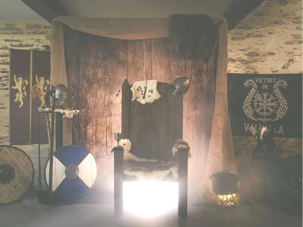 Une scène de décor médiéval avec un trône en bois, un drapeau avec des griffons et le mot 'Vikings' en arrière-plan, des accessoires de guerrier comme un bouclier et une épée.
