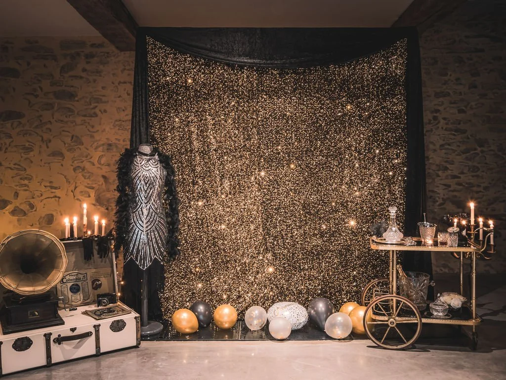 Décor de fête avec un fond noir scintillant, ballon dorés, argentés, et blancs, mannequin en costume de style Anneés 20, table avec des verres et bouteilles, et un gramophone vintage.