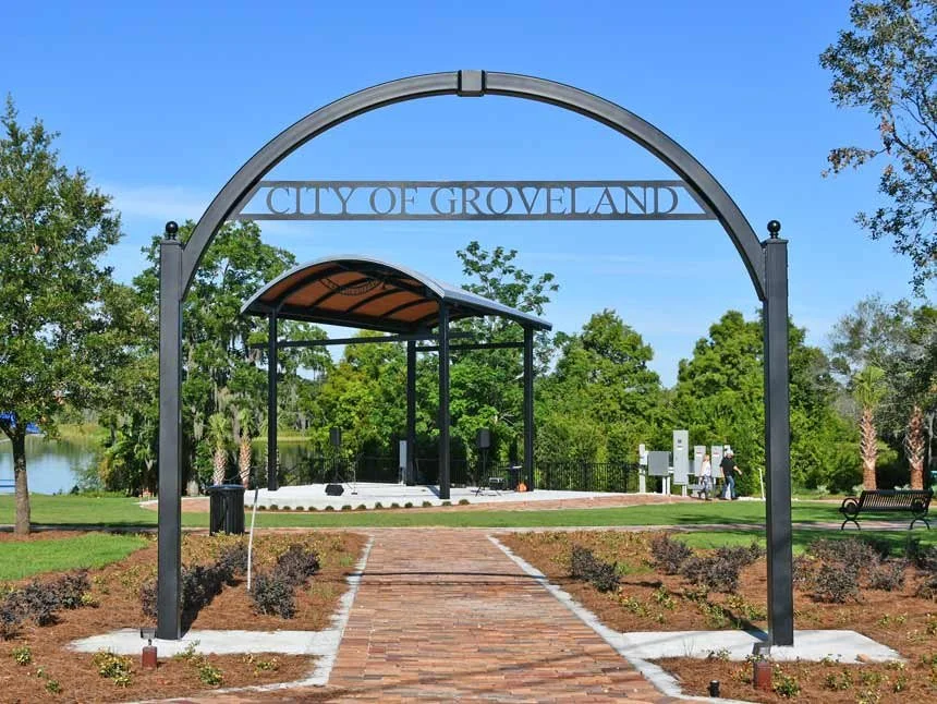Archway entrance with 'City of Groveland' sign over brick pathway leading to a park with a shelter, trees, and a pond in the background.