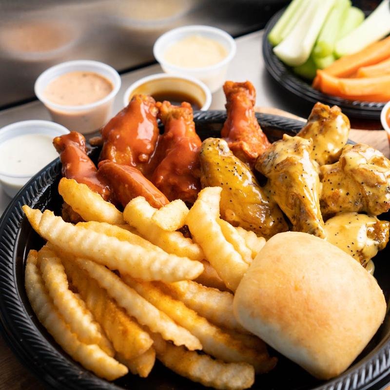 A black basket filled with fried chicken wings, crinkle-cut French fries, and a dinner roll, with small cups of dipping sauces in the background and a plate of celery and carrot sticks.