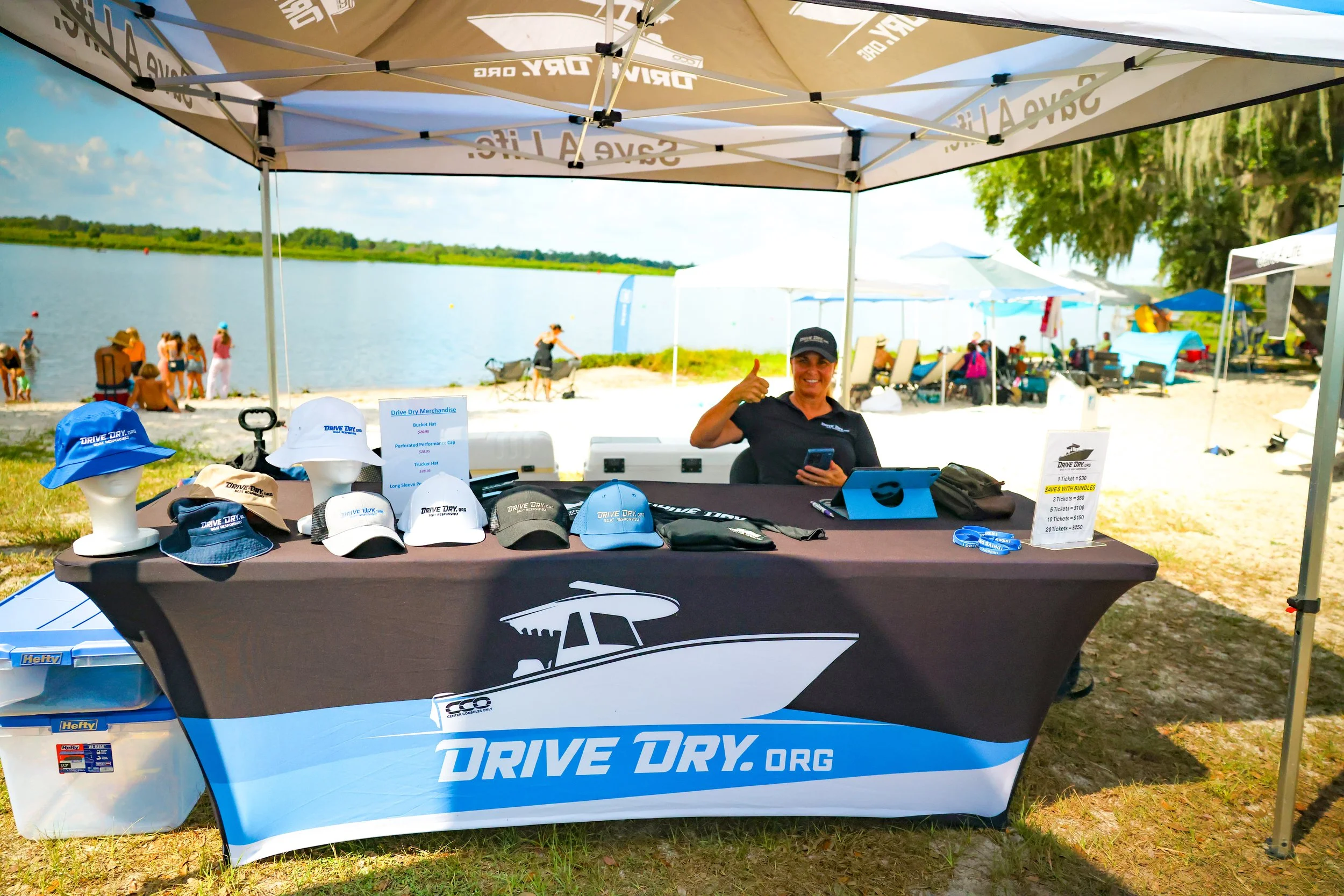 A smiling woman sitting at a booth with a Drive Dry.org banner, wearing a black shirt and cap, giving a thumbs-up, with caps and merchandise on the table, set up near a lake with people and tents in the background.
