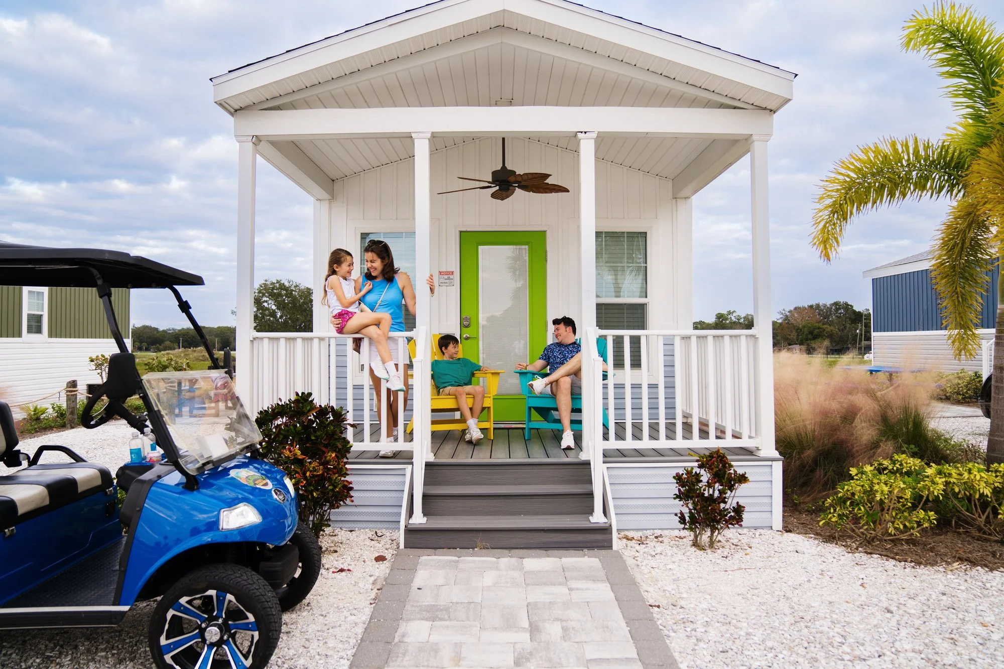 A family on a house porch with four people, a woman with two children and a man, sitting on chairs, talking and enjoying the outdoors. There is a golf cart parked in front, palm trees, and surrounding greenery.