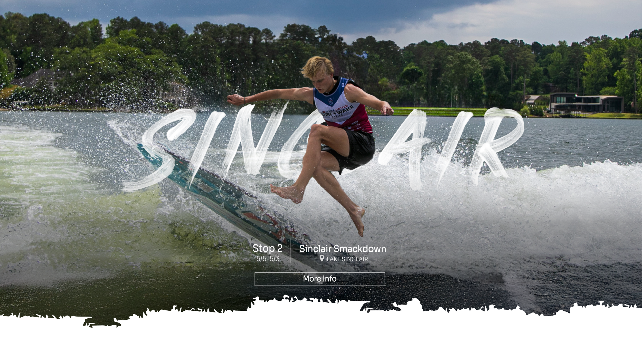 A person is wakeboarding on a lake, mid-air above the water, with trees and houses in the background.