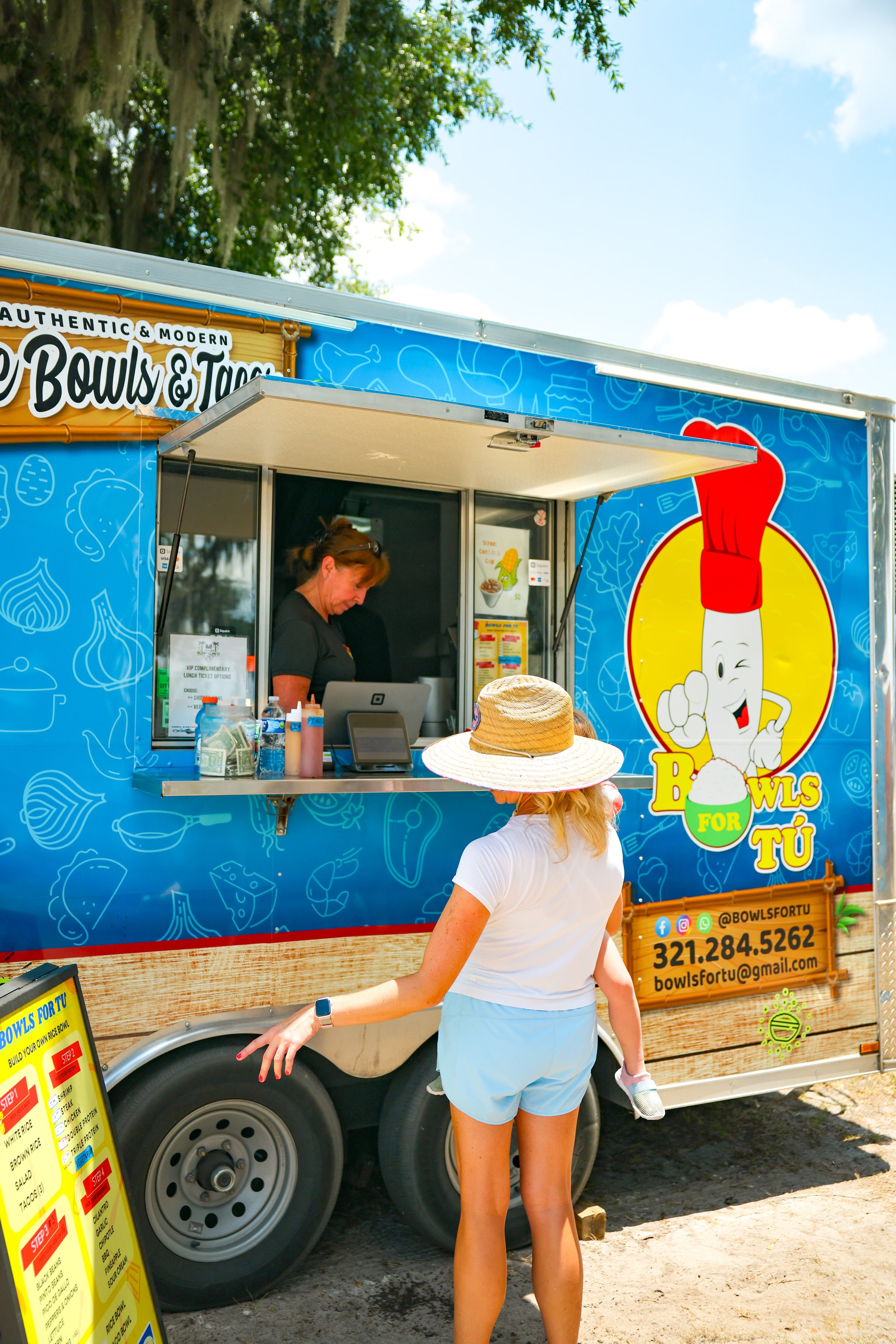A woman with a straw hat ordering food at a blue food truck called 'Bowls for Tu,' which serves authentic and modern bowls and tacos. The truck has a colorful logo of a cartoon bowl with a smiley face and a red chef's hat, and a menu sign outside.