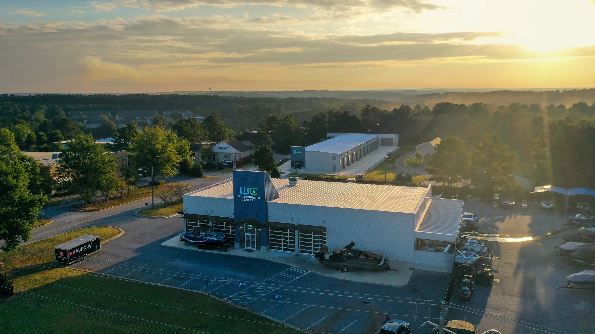An aerial view of Watersports Central retail store with boats displayed outside, parking lot, and surrounding trees at sunset.
