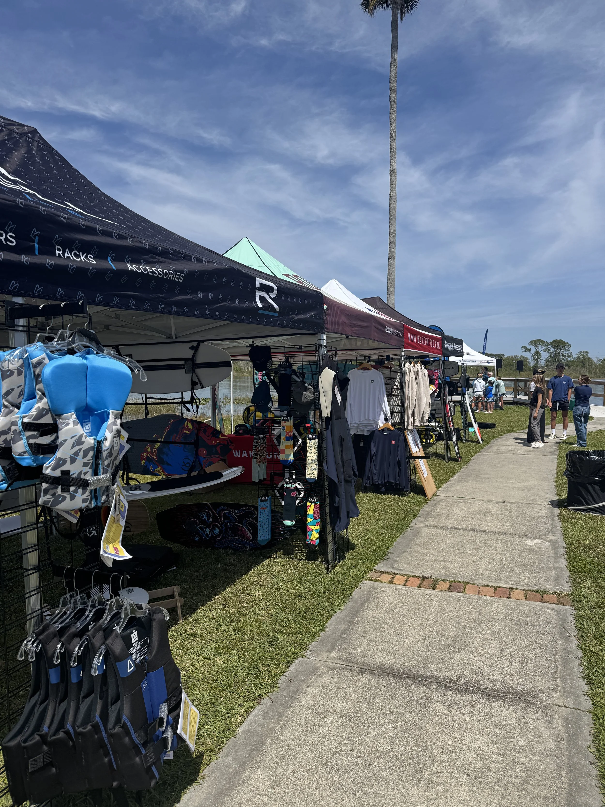 Outdoor marketplace with booths selling clothing, accessories, and sports gear under tents on a sunny day with a blue sky and a tall palm tree.