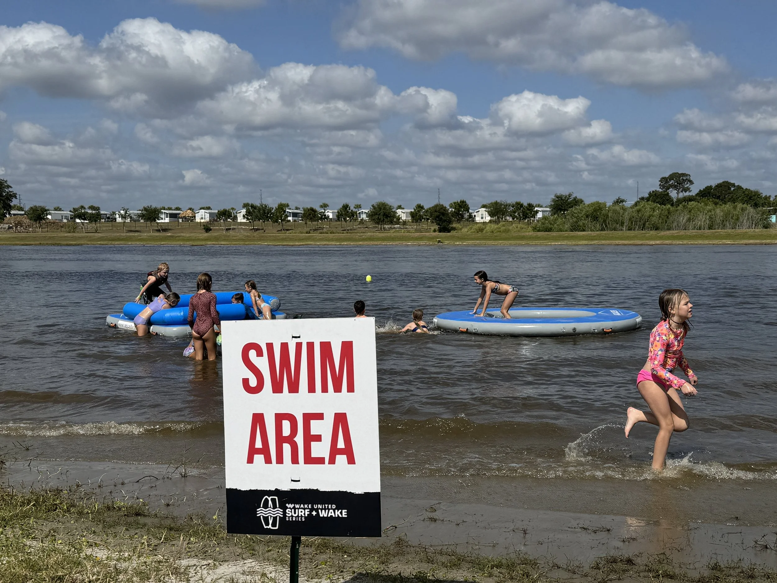 Children playing in a lake with paddle boards and an inflatable raft, under a cloudy sky, near a sign that says 'Swim Area'.