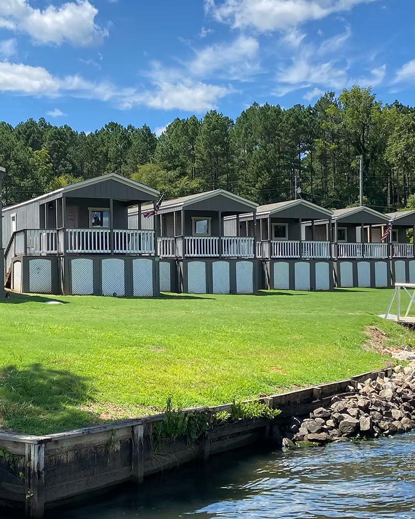 A row of gray houses with decks along a grassy shoreline, with water in the foreground and a wooded hill in the background under a partly cloudy sky.