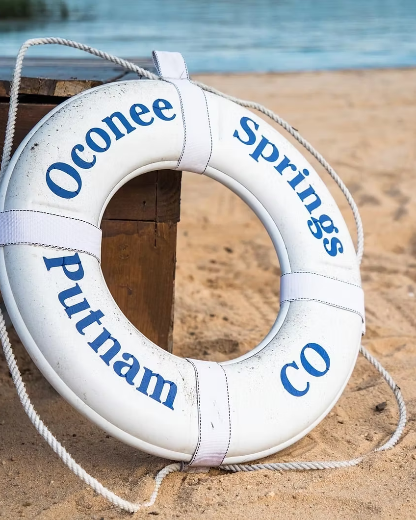 White lifebuoy with blue text reading 'Oconee Springs Putnam Co,' resting on a sandy beach near a wooden post, with water in the background.