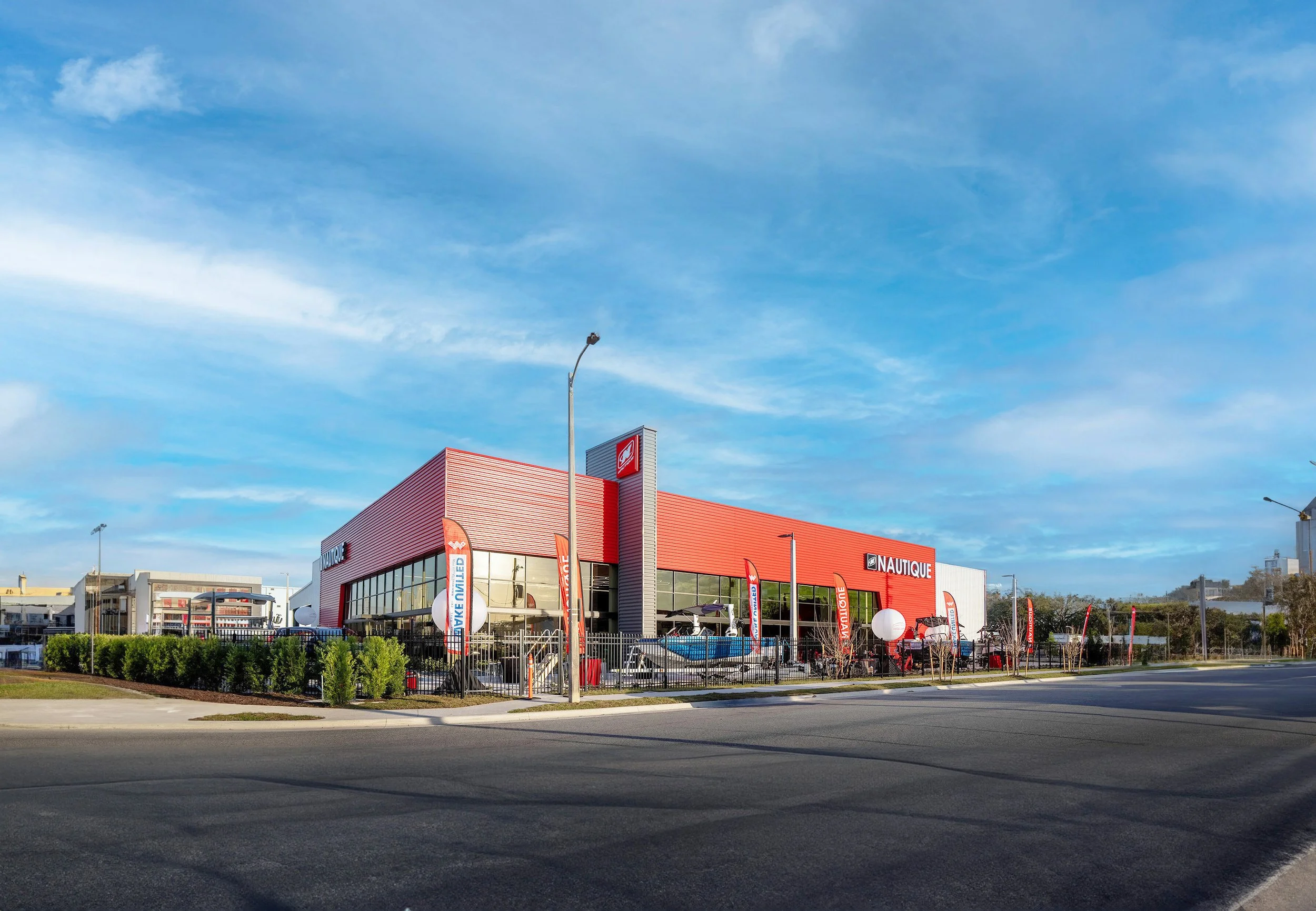 A red and gray boat dealership building with large glass windows, flags, and a boat display outside, under a partly cloudy sky.