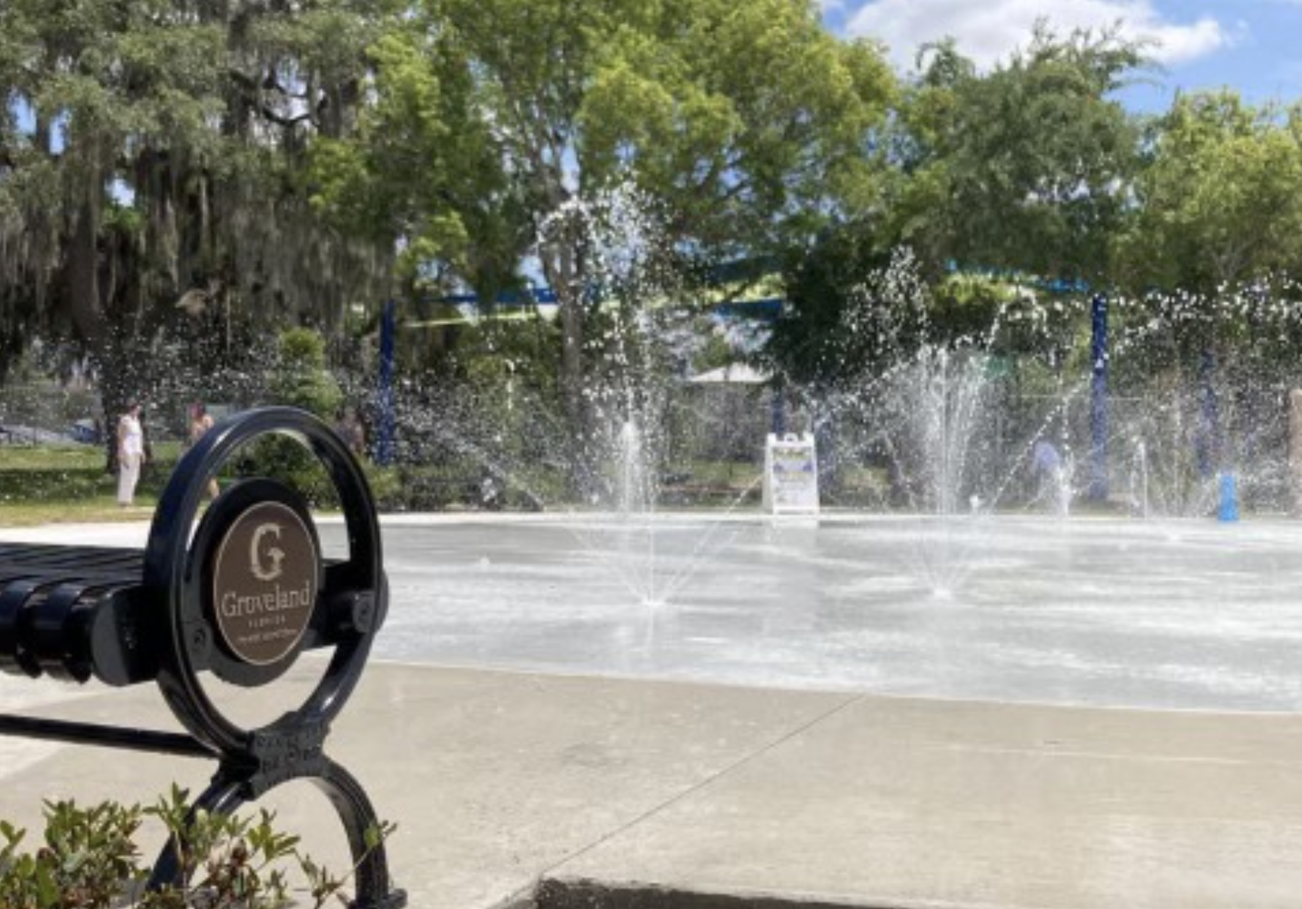 Fountain with water sprays in a park, playground area in the background, and trees with green leaves.
