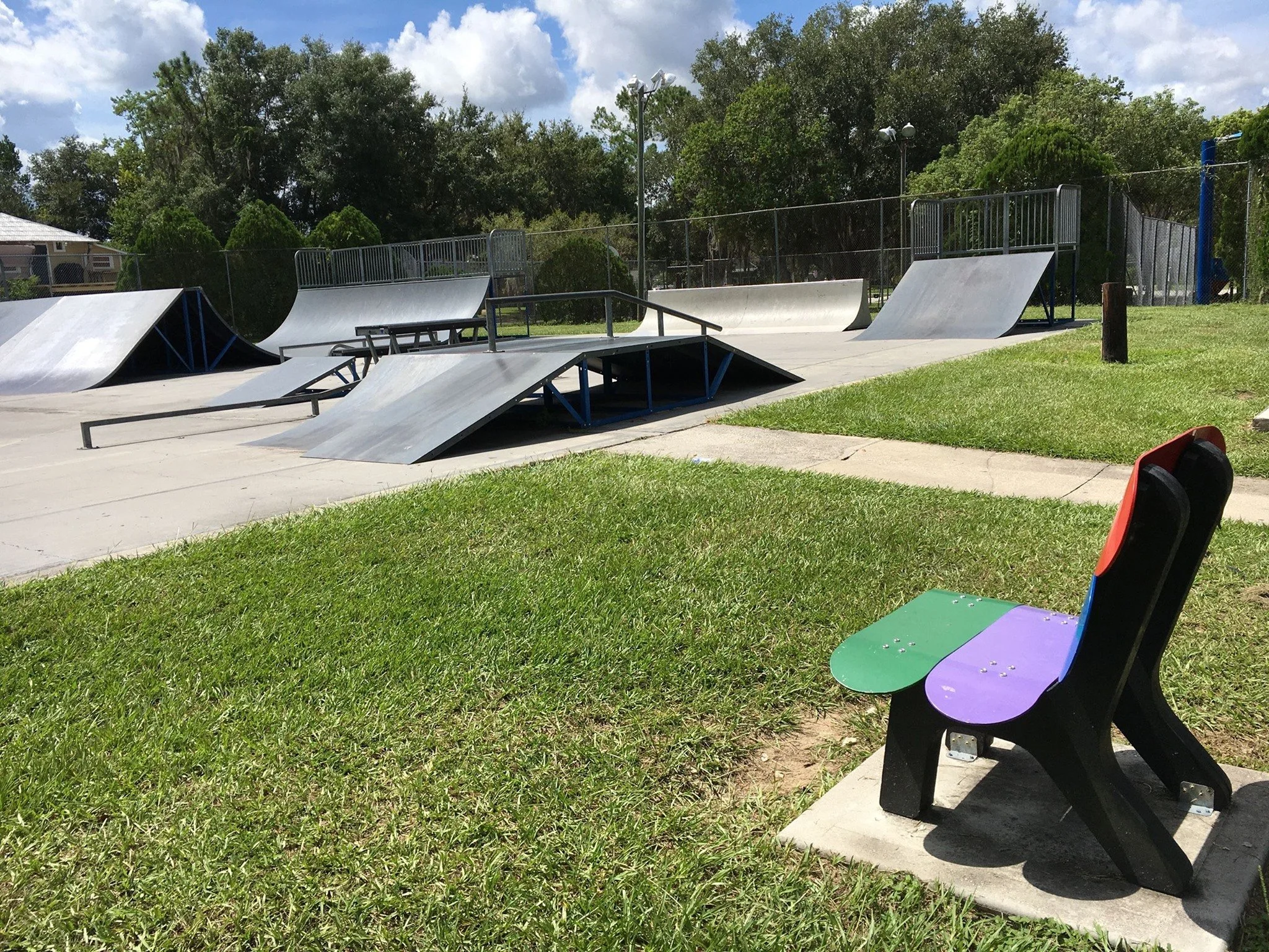 An outdoor skate park with multiple metal ramps and rails, surrounded by green grass and trees, under a partly cloudy sky, and a colorfully painted bench in the foreground.