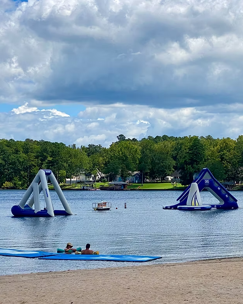 A lakeside scene with inflatable water slides in the water, a sandy beach area with two people on a paddleboard, green trees across the lake, and a cloudy sky overhead.