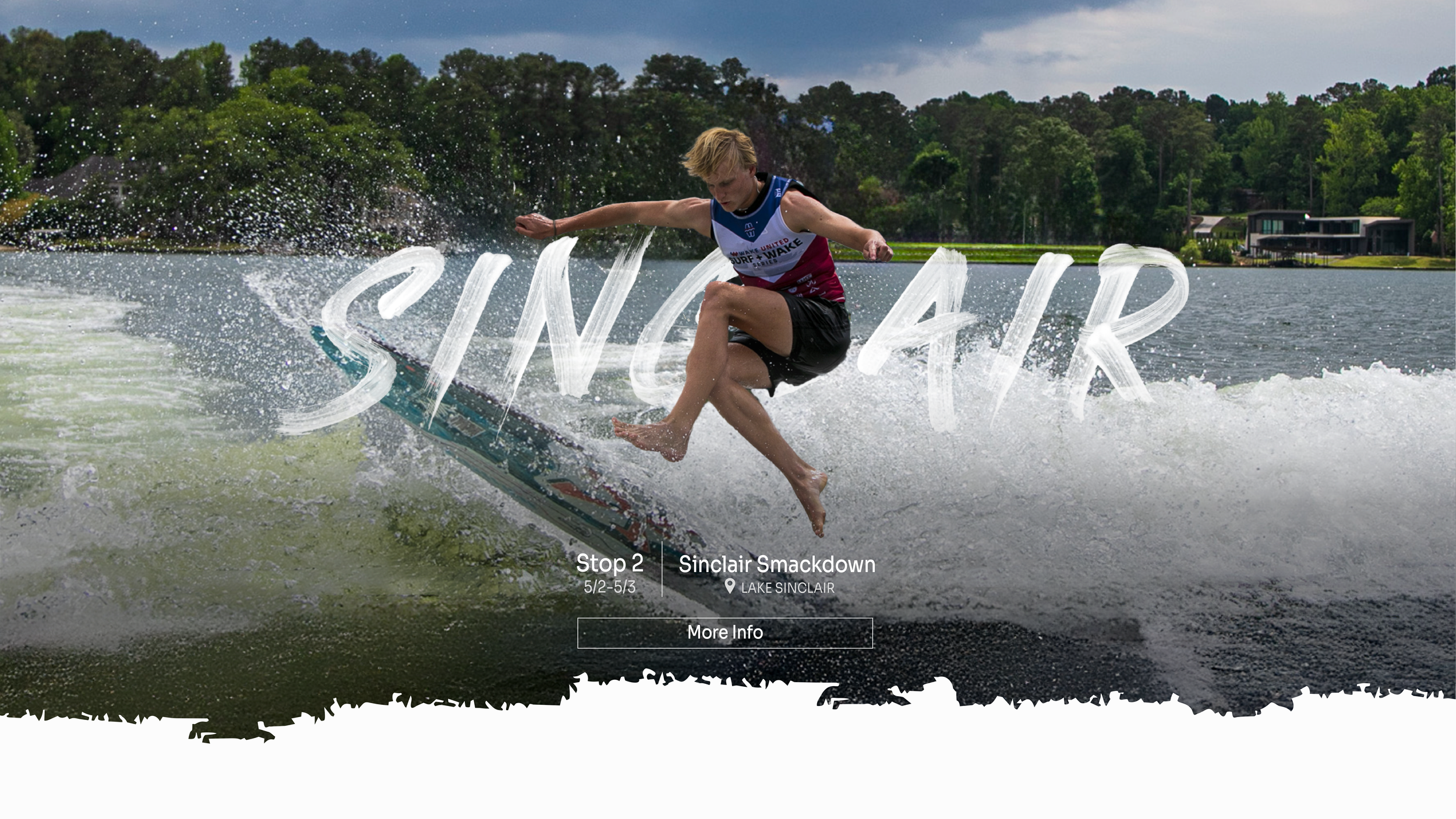 A person is wakeboarding on a lake, mid-air above the water, with trees and houses in the background.