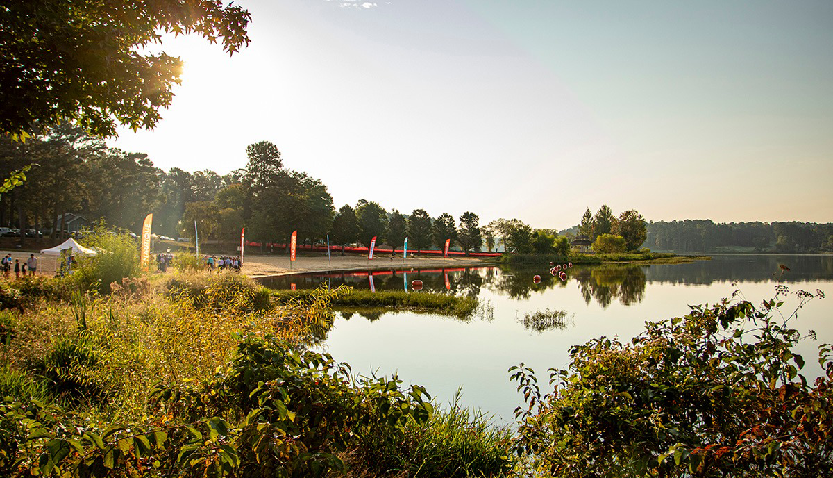 A lakeside scene with trees, a small beach area, people, and colorful flags, reflecting on calm water during sunset.