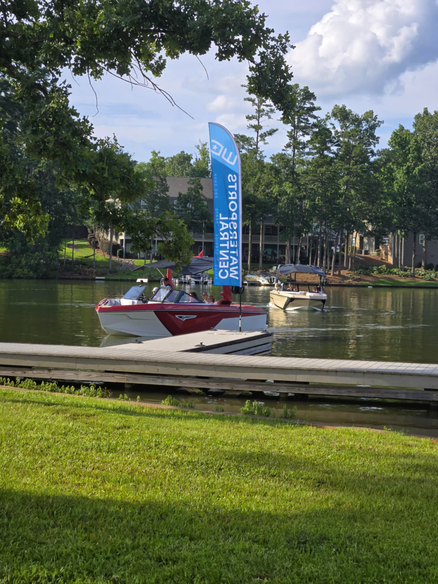 Boats docked by a pier on a lake with a blue flag saying 'SALES EVENT' and surrounded by trees and houses under a partly cloudy sky.